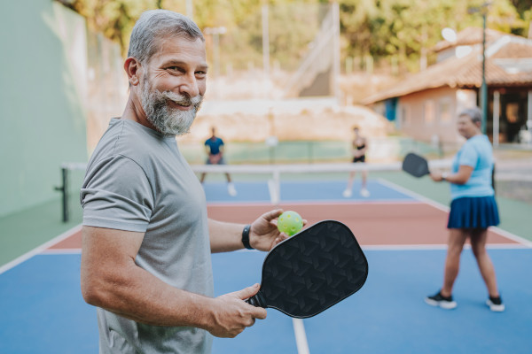 A man in the foreground of a pickleball court smiling at the camera during a match